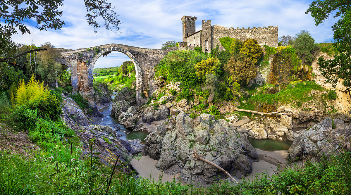 Vulci. Il Ponte dell’Abbadia, che supera il corso del sottostante fiume Fiora, e, sulla destra l’omonimo castello, il cui assetto attuale è frutto delle ristrutturazioni operate nel XII sec. e che, dal 1975, ospita il Museo Archeologico Nazionale.
